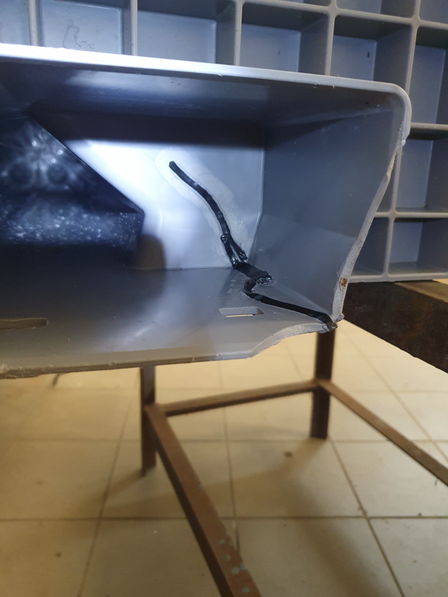 Szara plastikowa skrzynka z pęknięciem naprawionym czarnym klejem, widoczna na tle metalowego stelaża i kafelkowej podłogi.