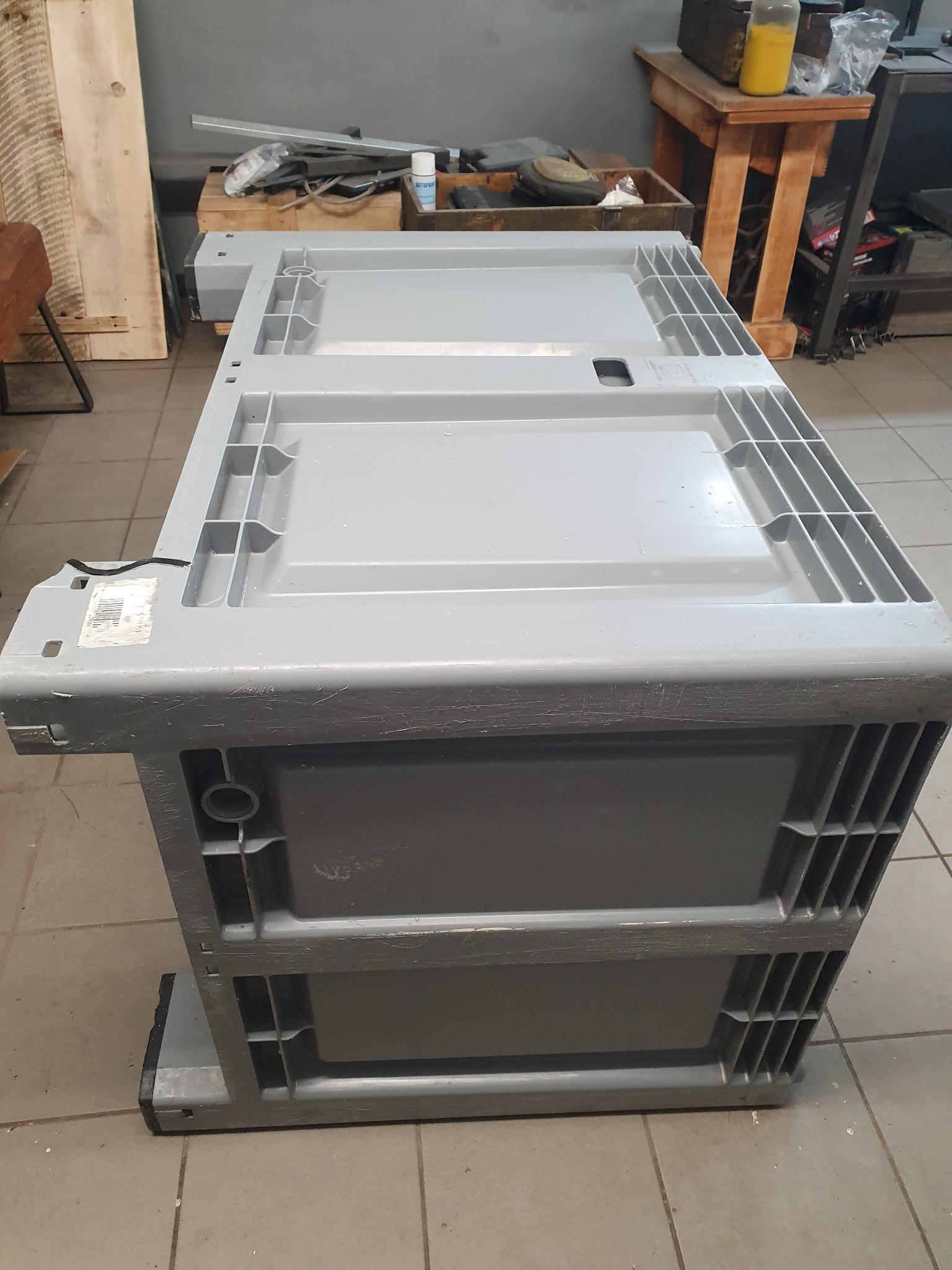 Szara, plastikowa skrzynia transportowa z ażurowymi ściankami, widziana z perspektywy żabiej, na tle warsztatu.