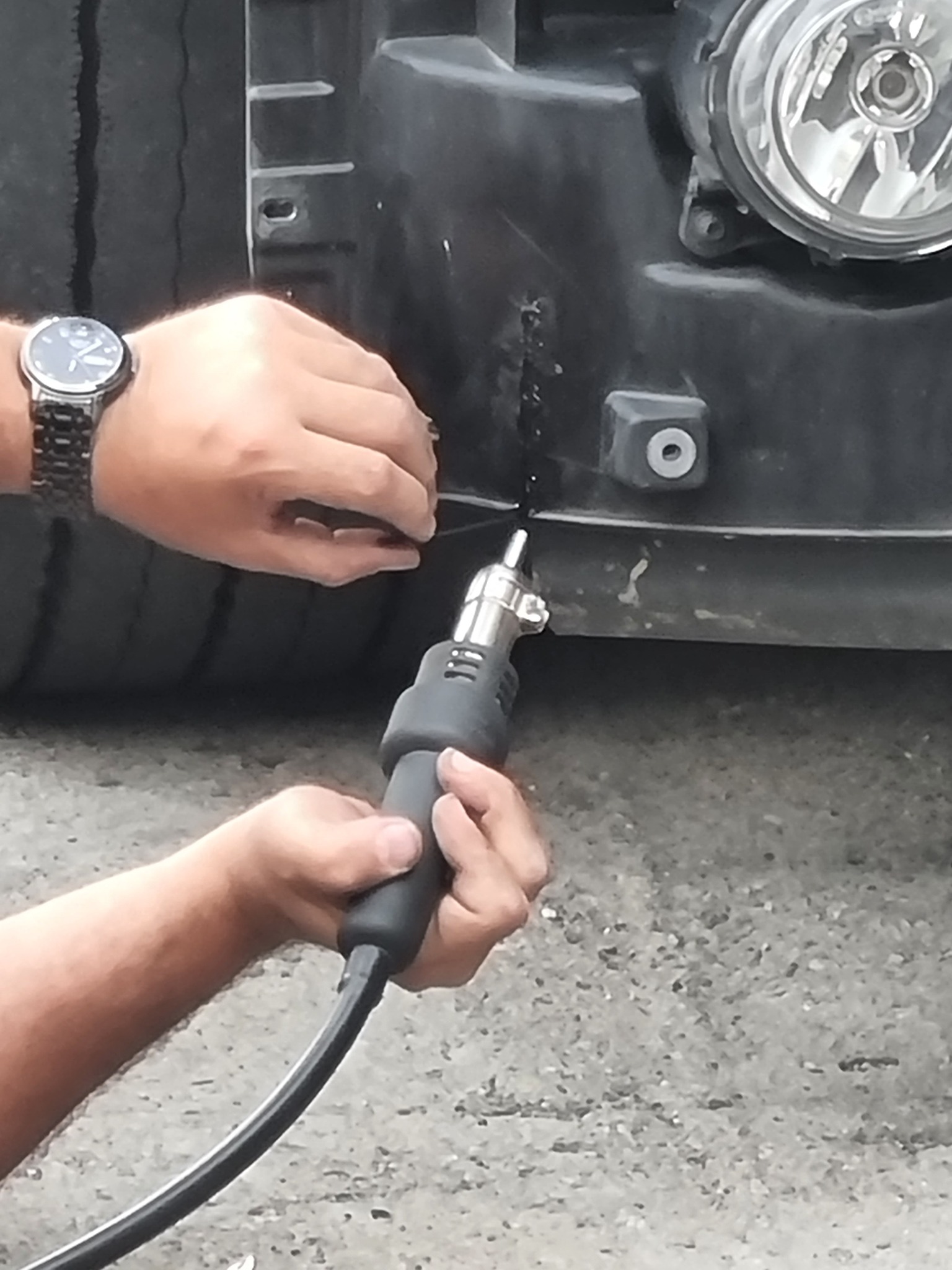 Naprawa plastikowego zderzaka samochodowego za pomocą opalarki; widoczne ręce, zegarek na nadgarstku, fragment opony i reflektora.