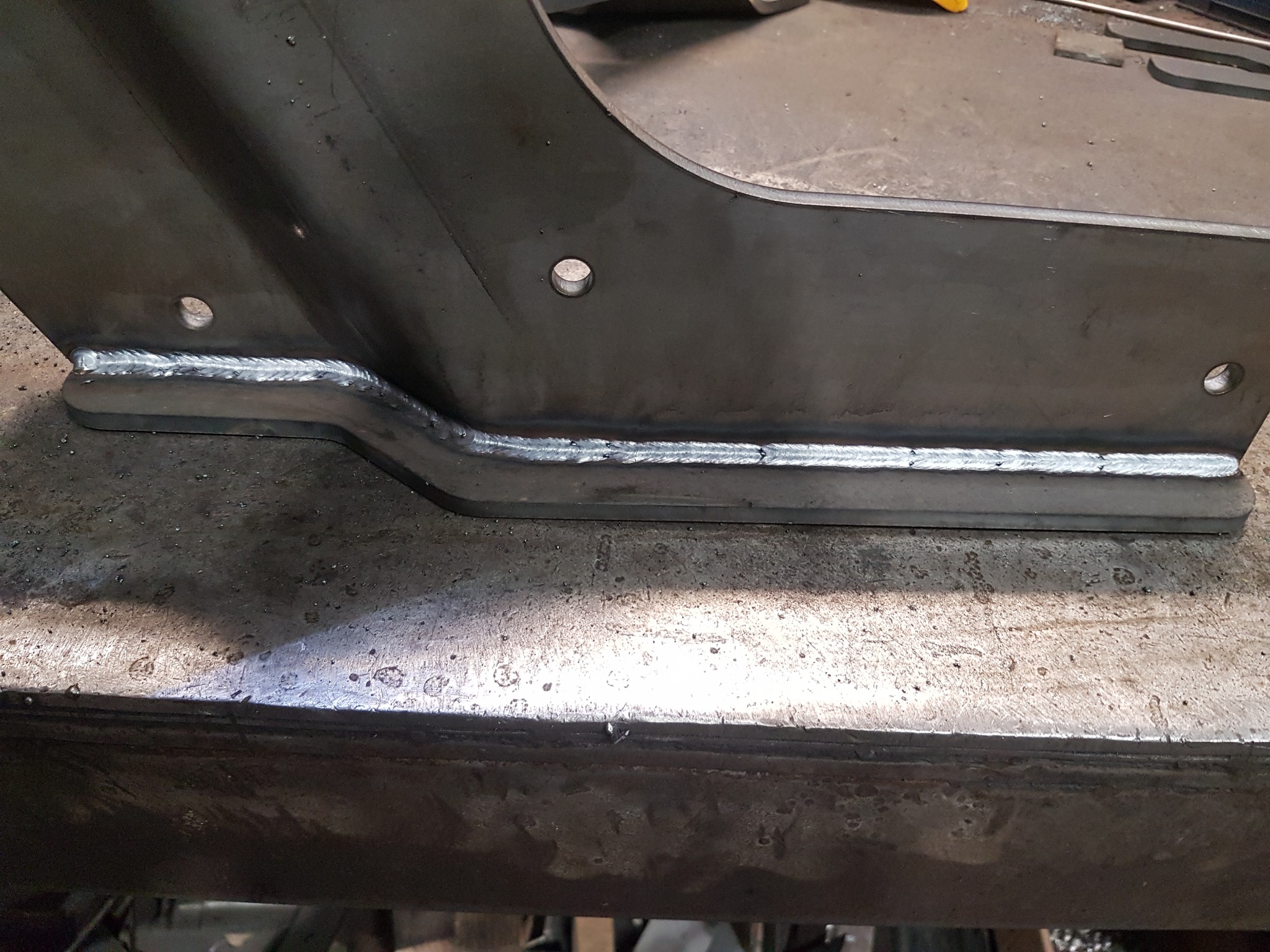 Metalowy element konstrukcyjny po spawaniu, widoczne spawy pachwinowe z charakterystycznym wzorem, trzy otwory montażowe, całość na warsztatowym stole.