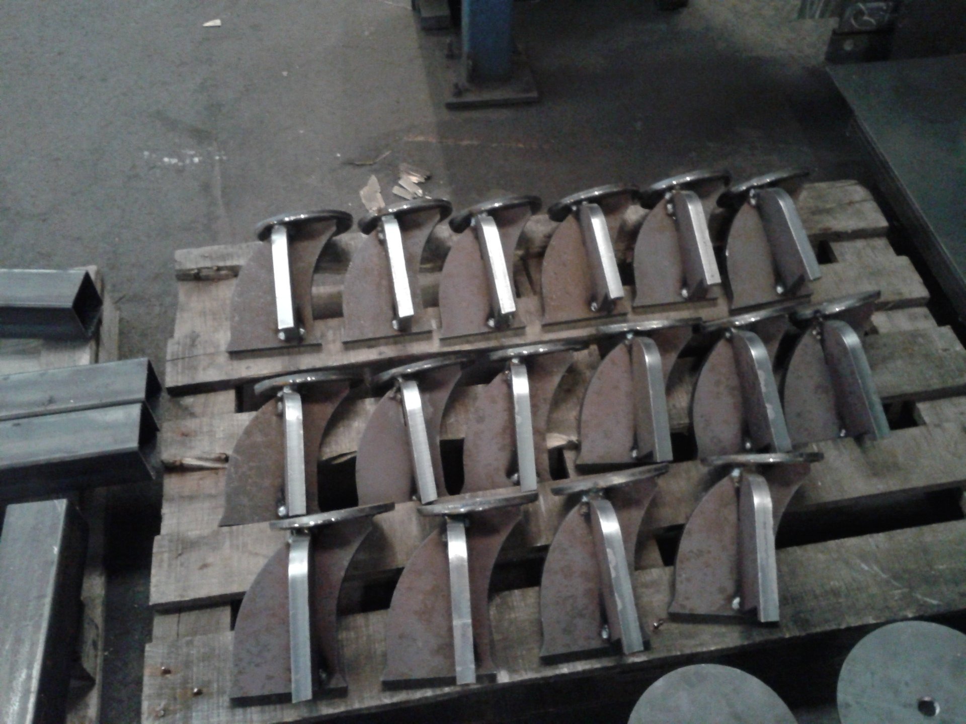 Rząd elementów metalowych, prawdopodobnie wsporników, ułożonych na drewnianej palecie w warsztacie, z widocznymi śladami obróbki.