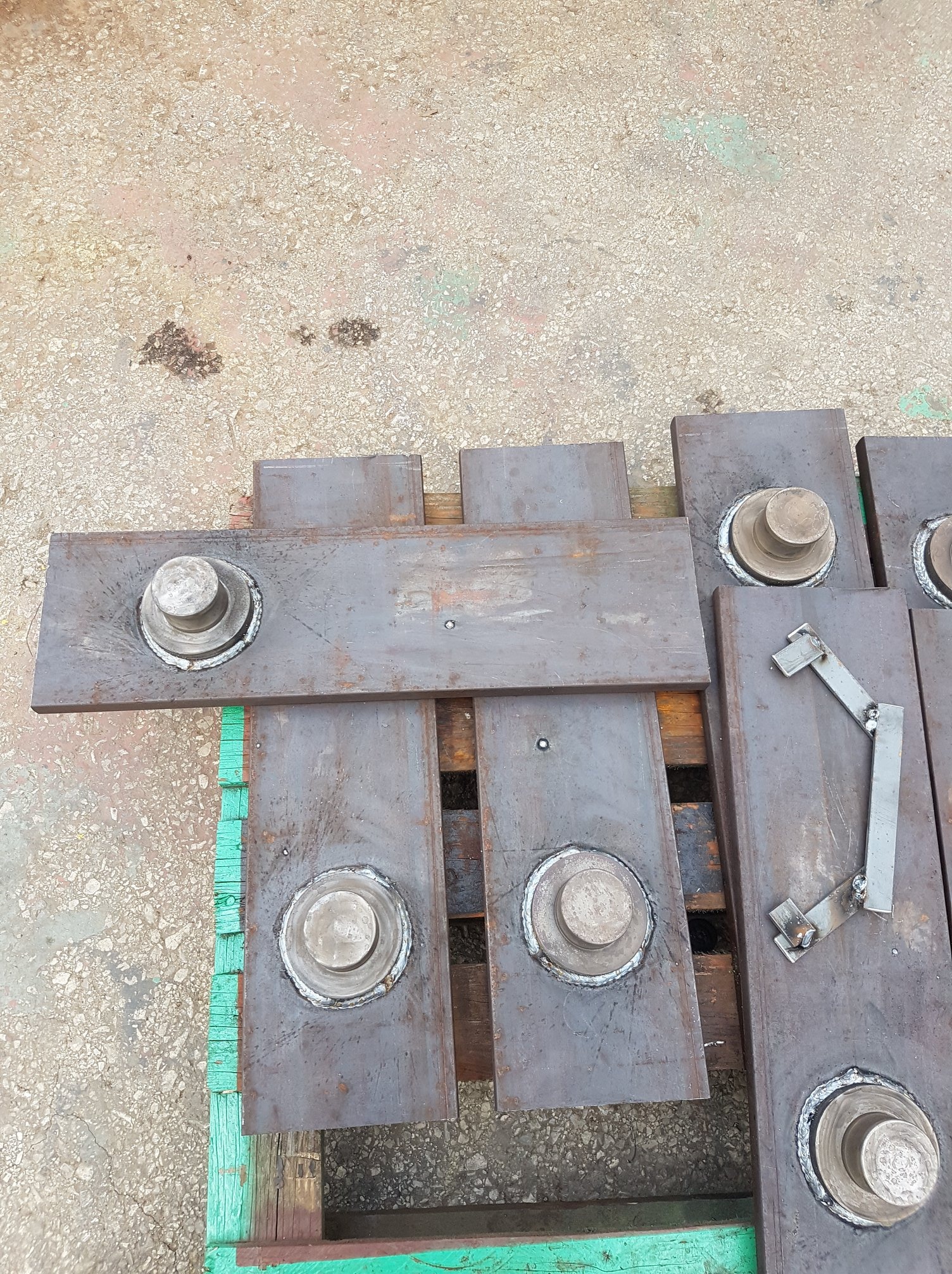 Metalowe płyty z przyspawanymi elementami, ułożone na drewnianej palecie pomalowanej na zielono; widoczne spoiny i ślady obróbki.