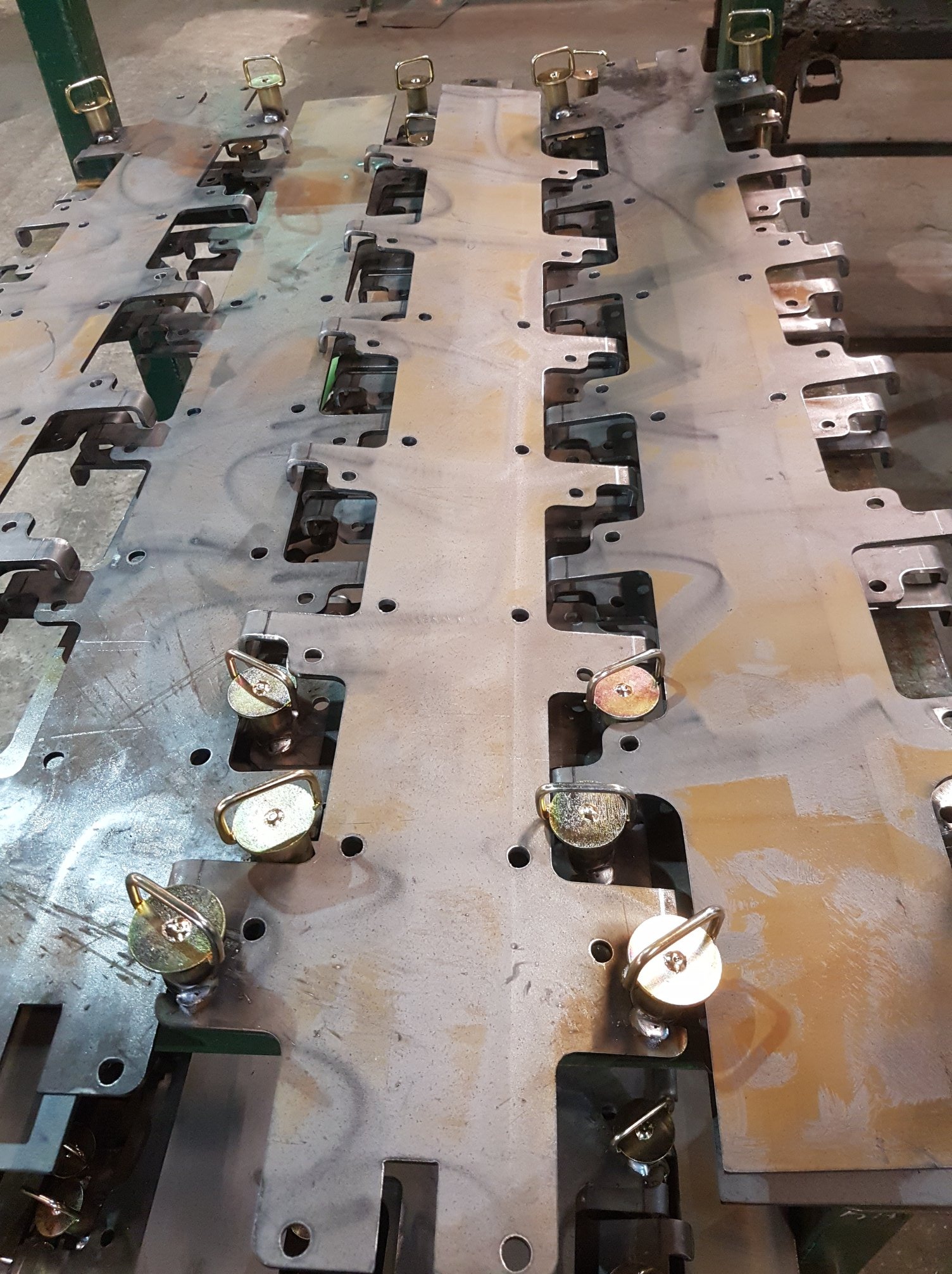 Metalowa płyta z wyciętymi otworami i przyspawanymi elementami mocującymi z uchwytami, widoczne ślady obróbki i delikatne przebarwienia na powierzchni.