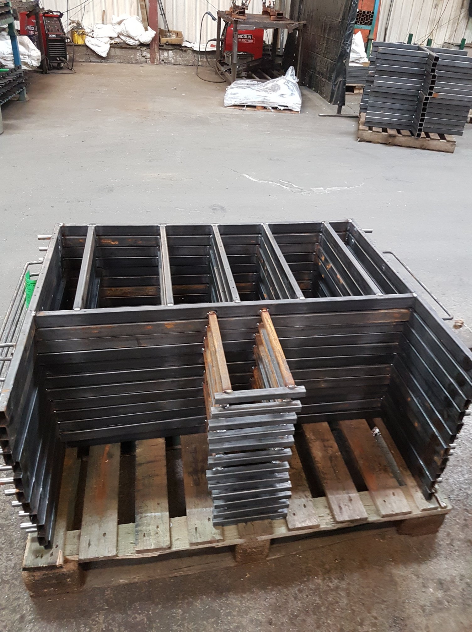 Stos metalowych, spawanych ram o prostokątnym przekroju ułożonych na drewnianej palecie w hali produkcyjnej.