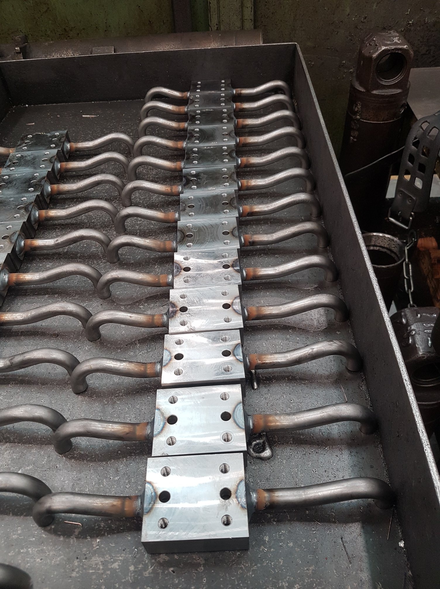 Rząd metalowych elementów z przyspawanymi rurkami w kształcie litery U, ułożonych w metalowej skrzyni; widoczne ślady po spawaniu.
