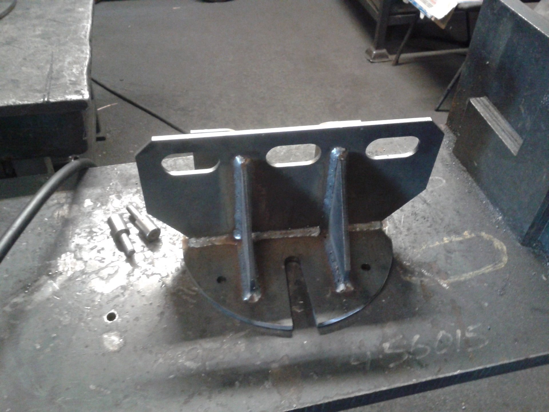 Metalowy wspornik spawany z blachy i prętów, widoczne spawy, leżący na metalowym stole warsztatowym obok narzędzi