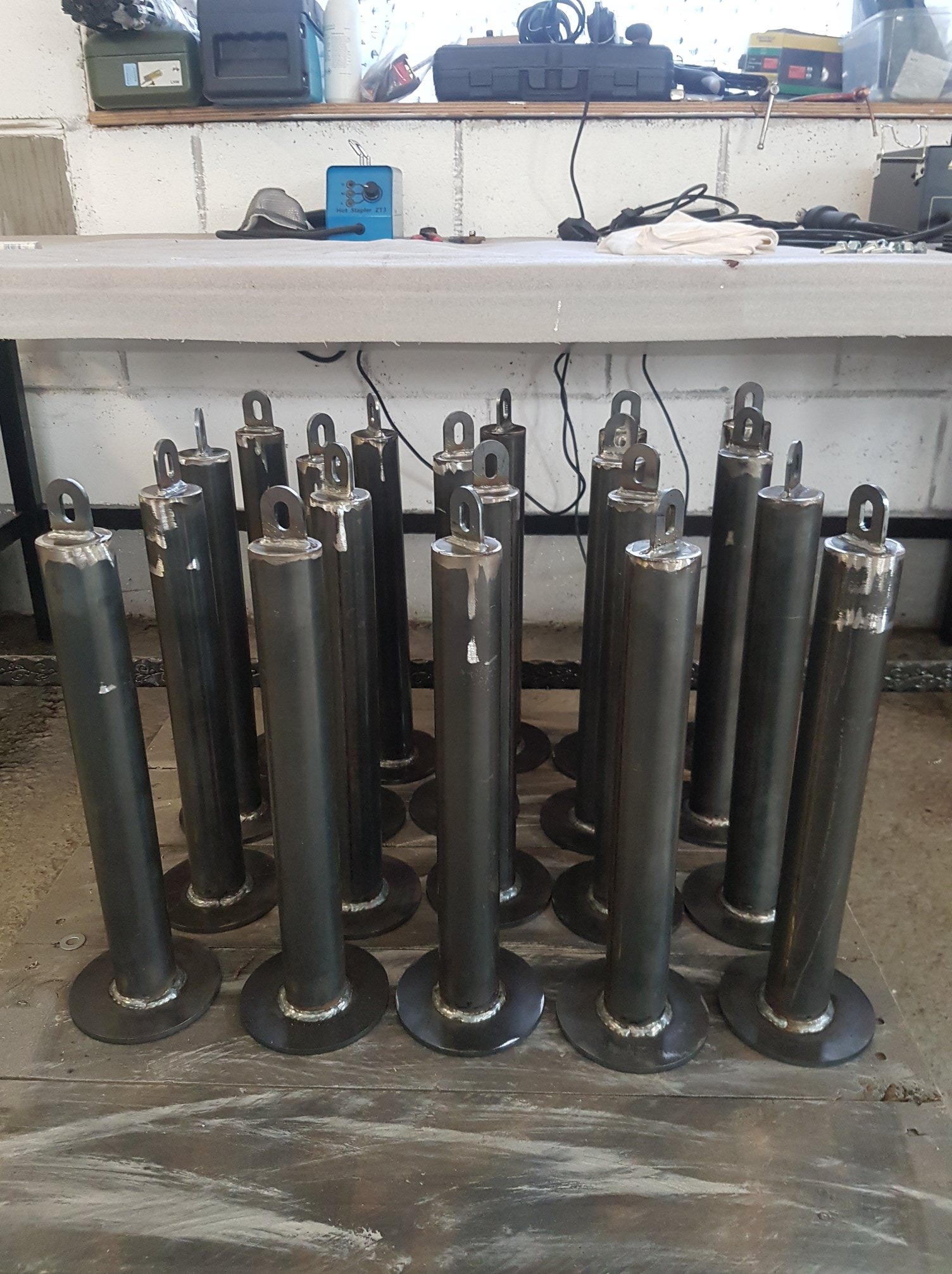 Seria spawanych, metalowych cylindrów z okrągłą podstawą i uchwytem, stojących na podłodze warsztatu, widoczne ślady obróbki na górnej krawędzi niektórych elementów.