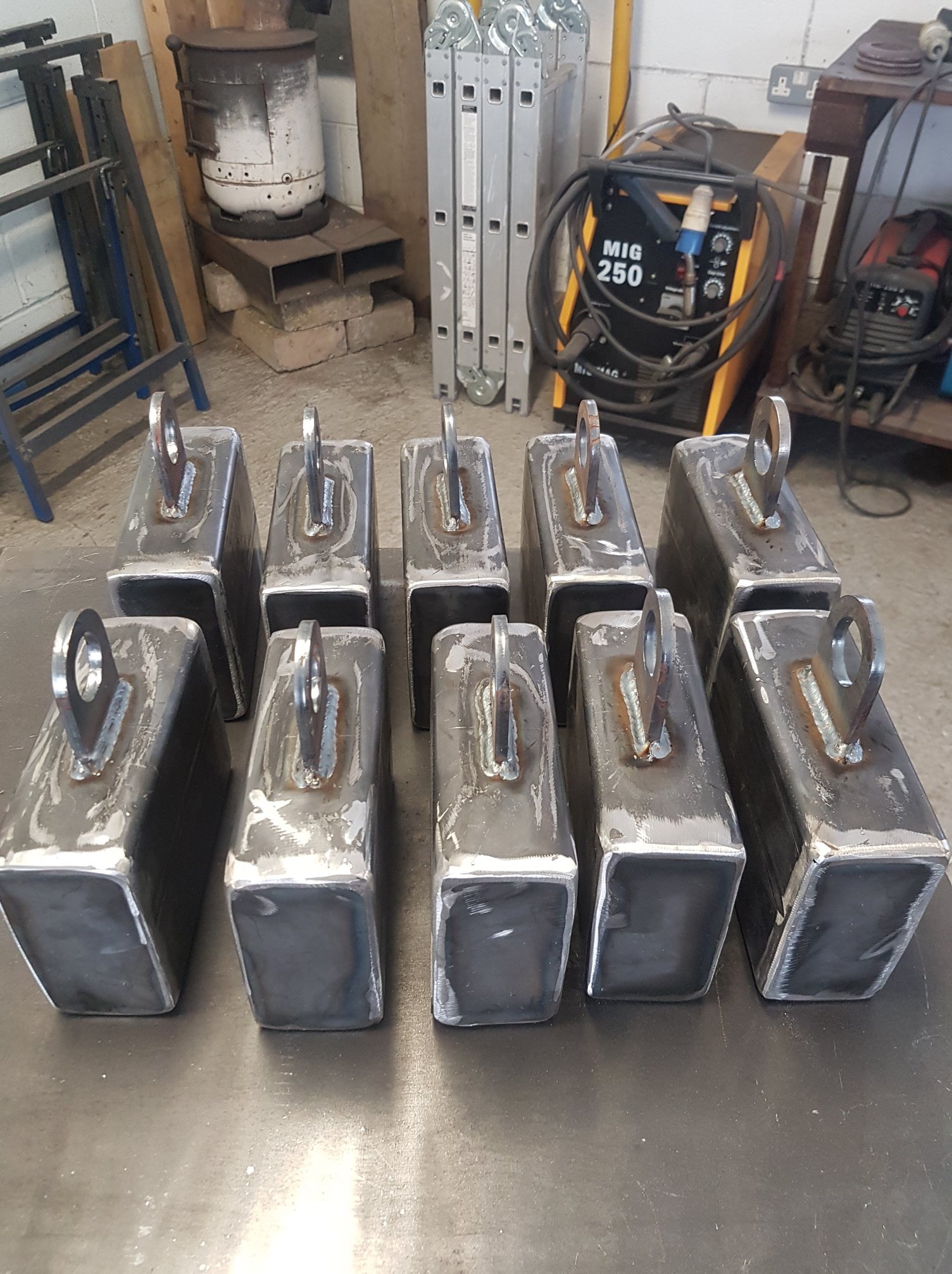 Partia spawanych, surowych, stalowych elementów w kształcie prostopadłościanów z przyspawanymi uchwytami na płaskiej powierzchni roboczej, w tle spawarka MIG 250 i drabina.