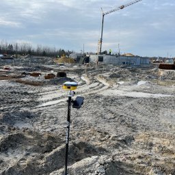 GEODETA - Żółty odbiornik GNSS marki E-Survey na czarnym tyczce z napisem FORGED wbity w błotnisty teren budowy, w tle fundamenty budynku i żuraw budowlany.