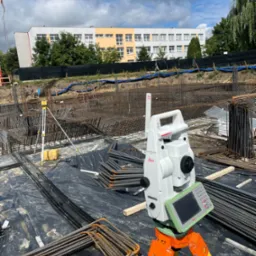 Tacheometr Leica na pomarańczowo-żółtym statywie, ustawiony na czarnej folii z rozłożonym zbrojeniem budowlanym, w tle budynek i pochmurne niebo.