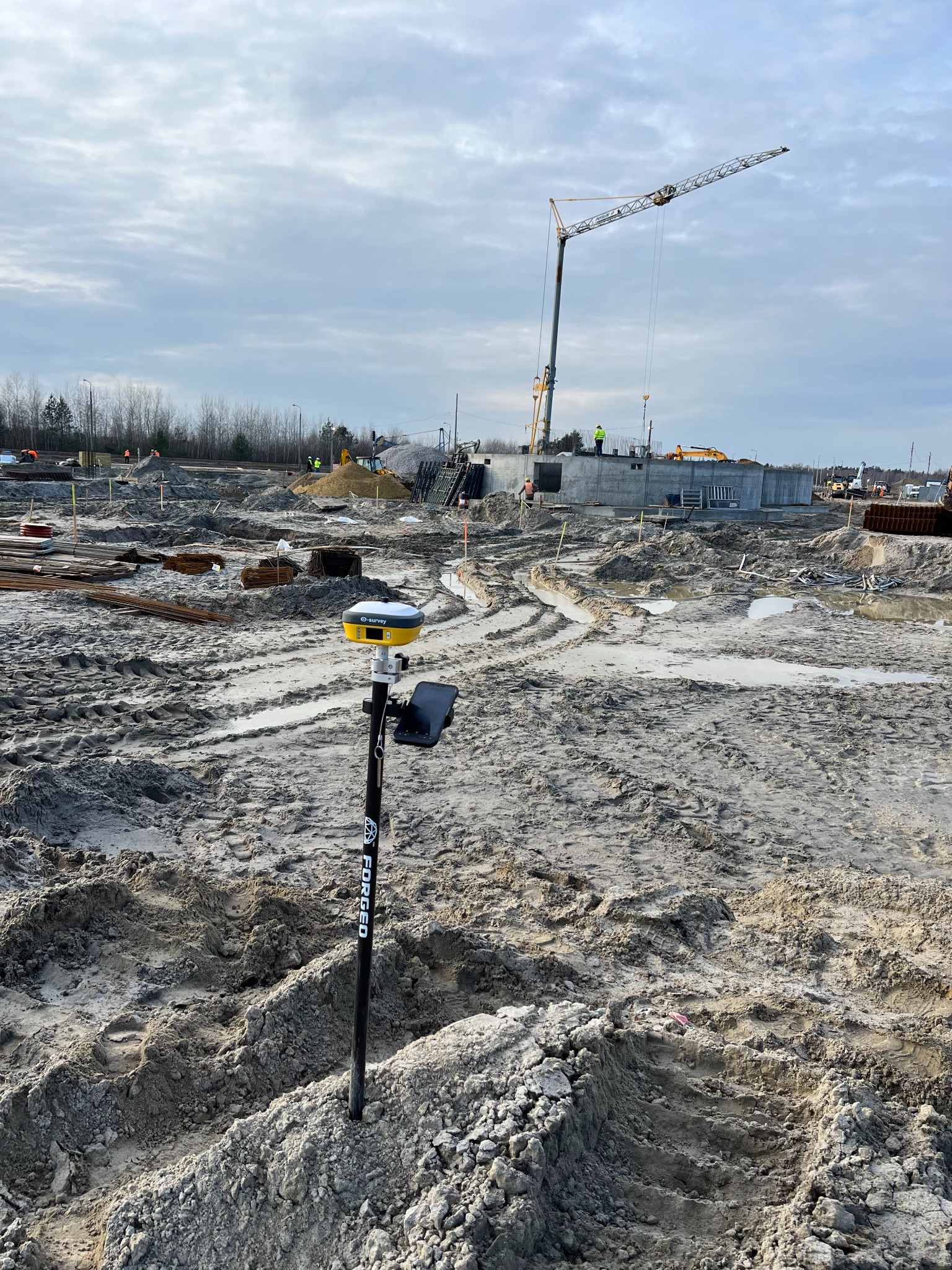 Żółty odbiornik GNSS marki E-Survey na czarnym tyczce z napisem FORGED wbity w błotnisty teren budowy, w tle fundamenty budynku i żuraw budowlany.