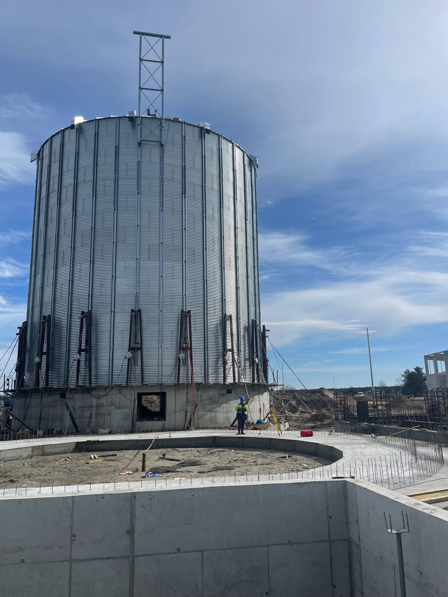 Budowa silosu zbożowego z metalowym płaszczem, widoczne betonowe fundamenty i pracownik w odblaskowej kamizelce na placu budowy, niebieskie niebo z chmurami w tle.