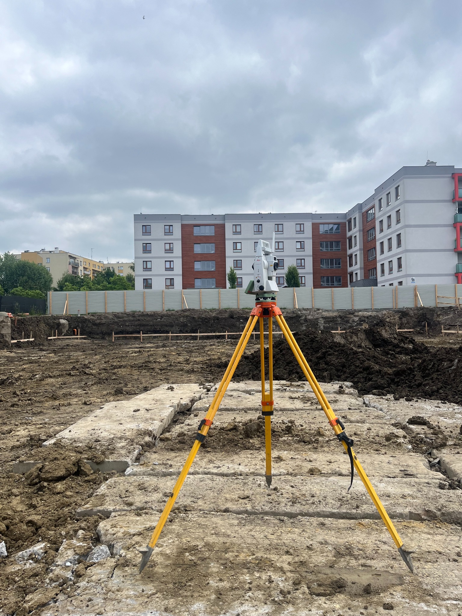 Tachimetr na żółtym statywie ustawiony na placu budowy z widocznymi fundamentami i budynkiem mieszkalnym w tle, pochmurne niebo.