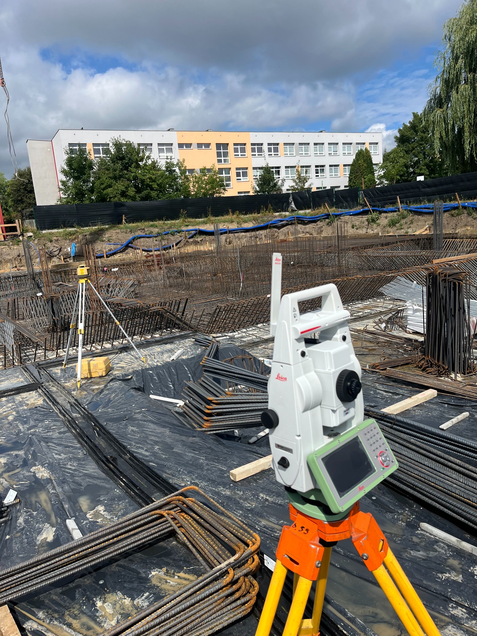 Tacheometr Leica na pomarańczowo-żółtym statywie, ustawiony na czarnej folii z rozłożonym zbrojeniem budowlanym, w tle budynek i pochmurne niebo.