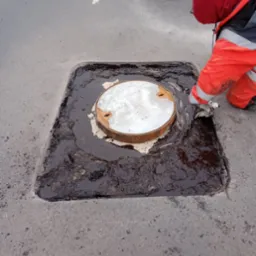 Pracownik w pomarańczowym kombinezonie czyści okolice studzienki kanalizacyjnej z brudną wodą na tle ubijarki i pachołka drogowego.