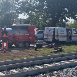 Pracownicy w pomarańczowych kombinezonach naprawiają nawierzchnię drogi przy użyciu zagęszczarki w pobliżu torów tramwajowych, widoczny samochód serwisowy z napisem POGOTOWIE WOD-KAN oraz maszyna...
