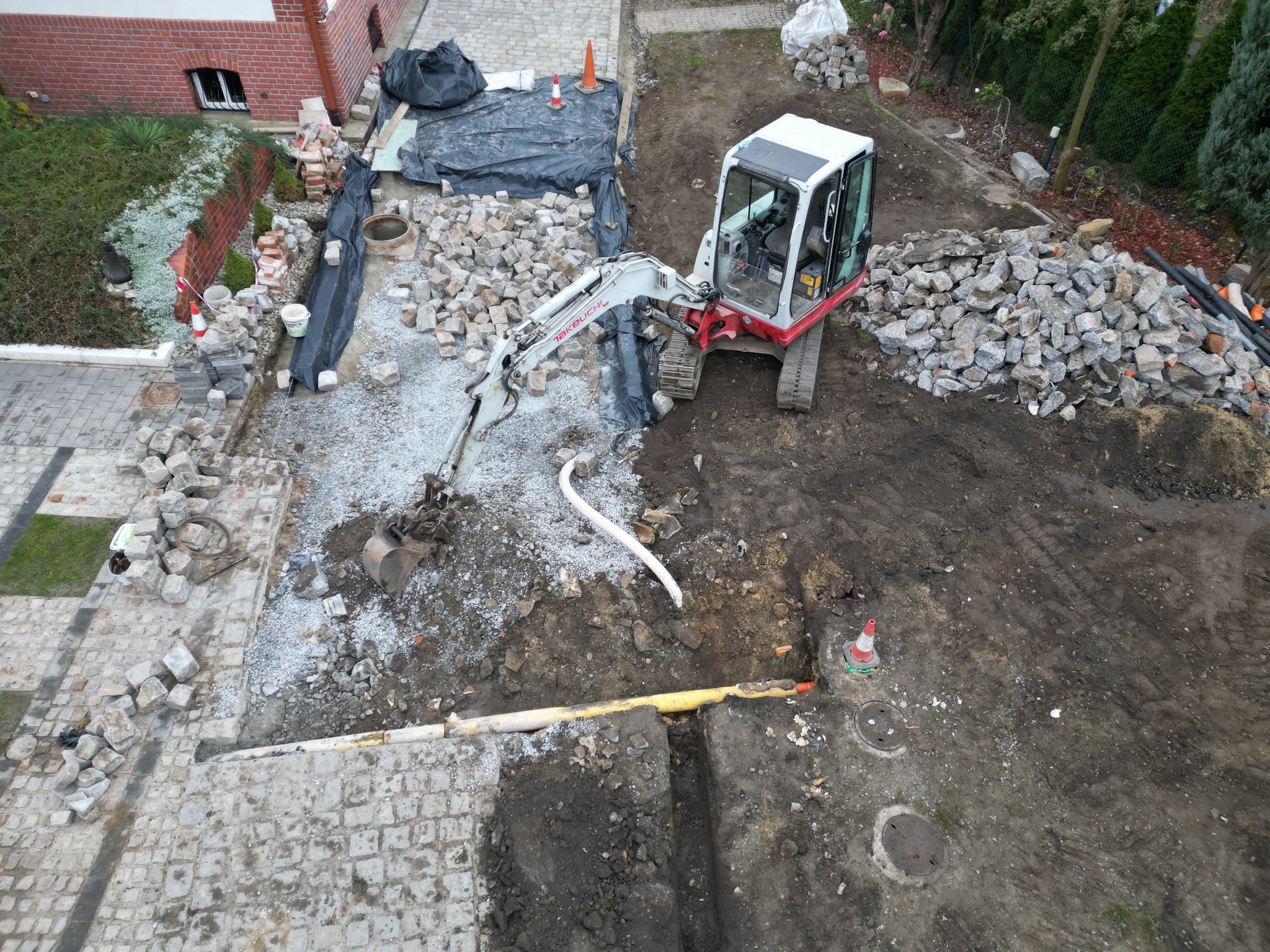 Widok z góry na plac budowy z mini koparką Takeuchi, przygotowany teren pod kostkę brukową, widoczne rury i studzienki kanalizacyjne oraz sterty kamieni