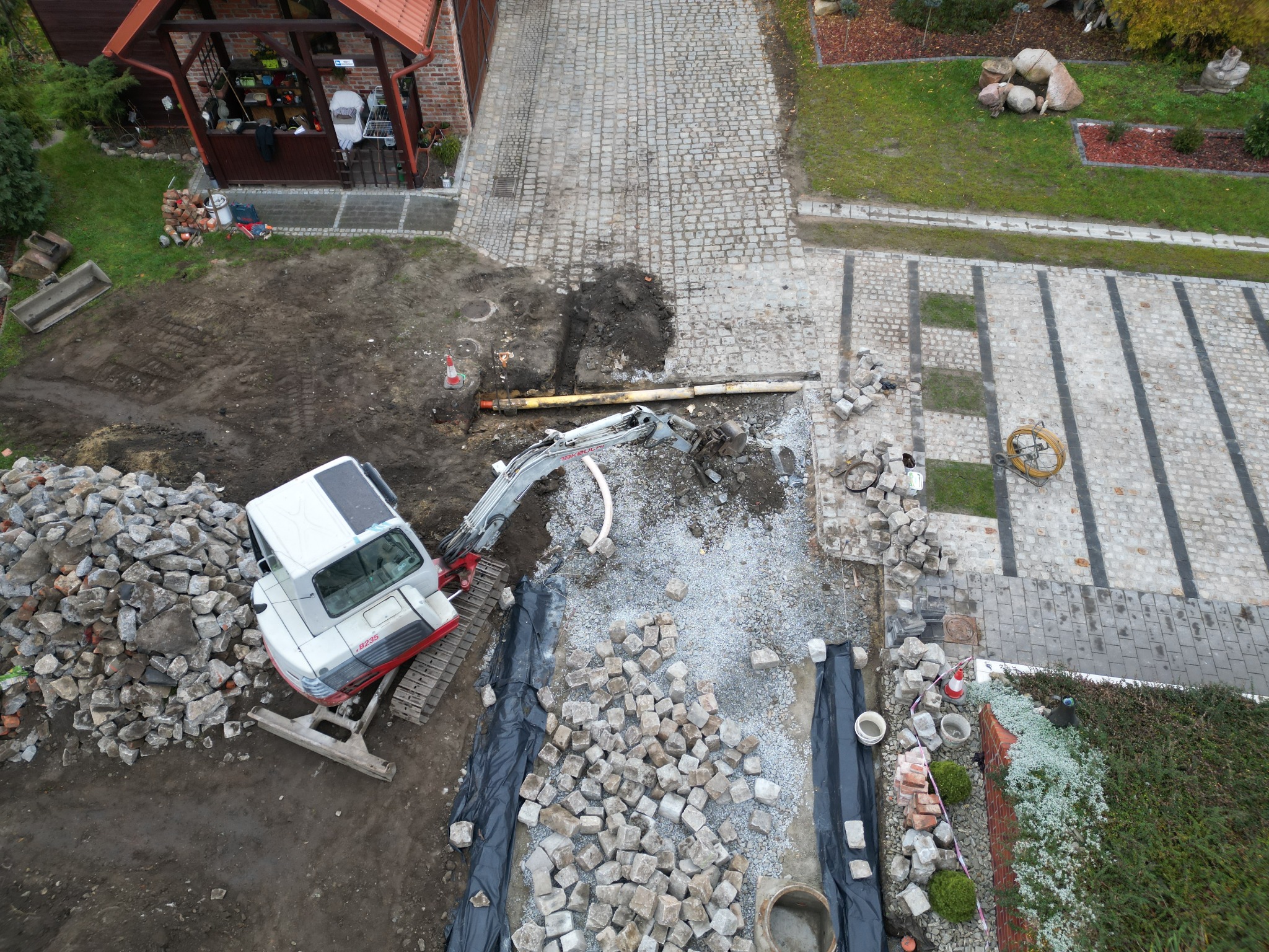 Zdjęcie z góry przedstawia wykop pod rury, z widocznym minikoparką, stertą kamieni i fragmentem ułożonej kostki brukowej. Rury leżą w wykopie obok siebie. Widoczny również fragment posesji...