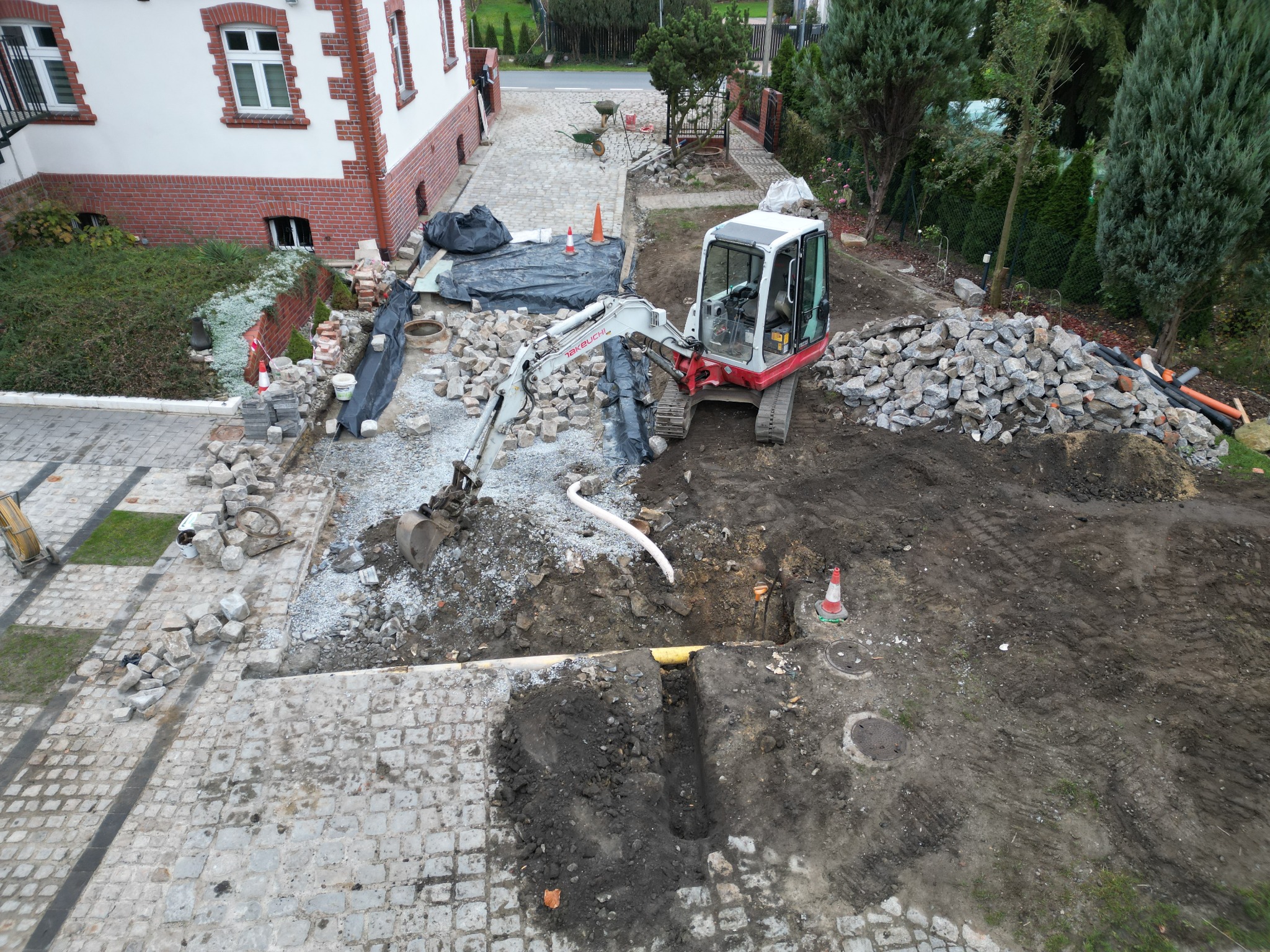 Mini koparka Takeuchi przy wykopie na posesji z kostki brukowej, widoczne rury, studzienki i przygotowane kamienie, dom w tle.