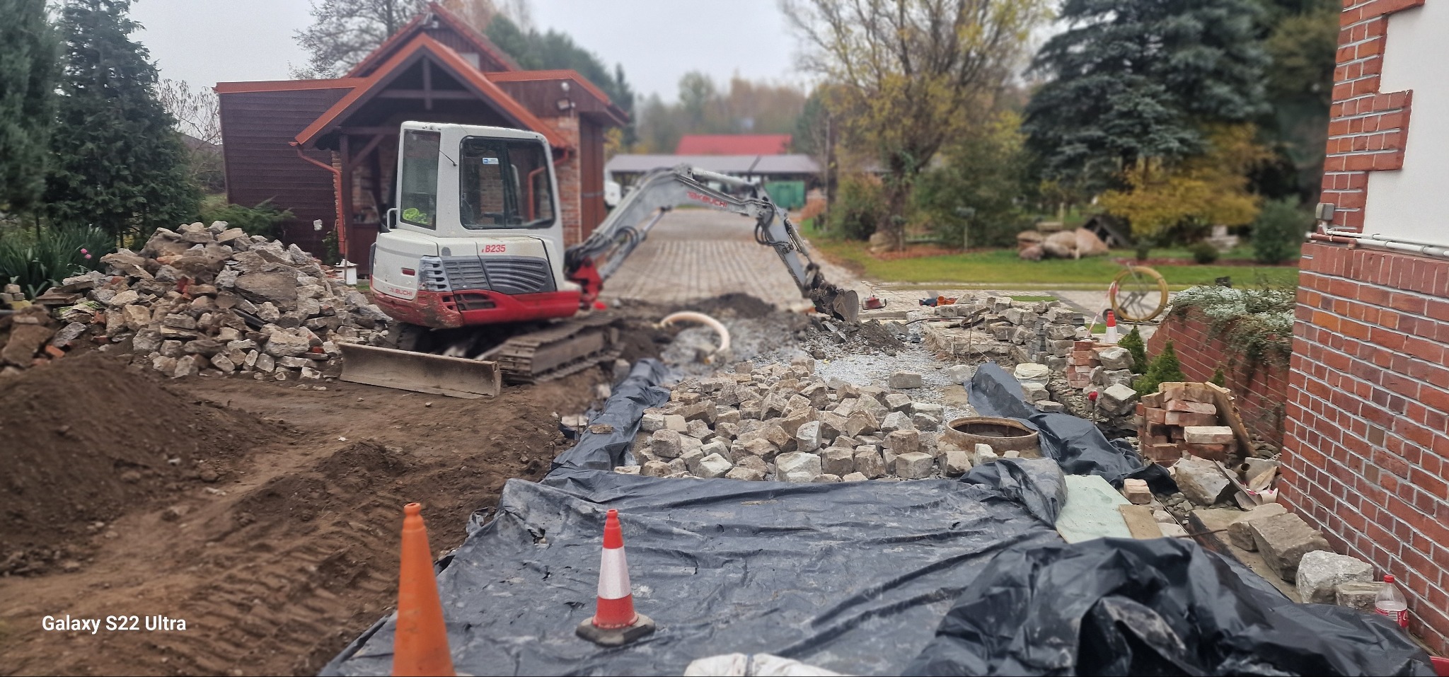 Mini koparka TAKEUCHI B235 w trakcie prac ziemnych związanych z instalacją rur na posesji, widoczne wykopane kamienie, zabezpieczenia i rury, w tle dom z brązową elewacją.