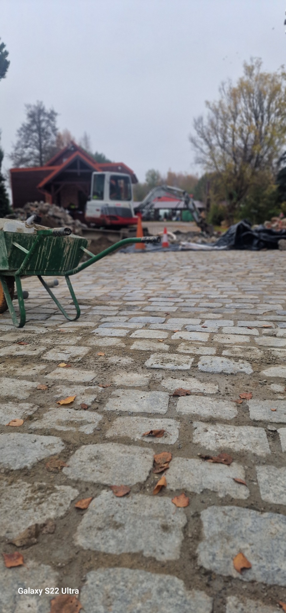 Ułożona kostka brukowa na pierwszym planie, w tle wykop z dwoma mini koparkami i zielona taczka z ziemią, jesienne liście na kostce, pochmurny dzień.