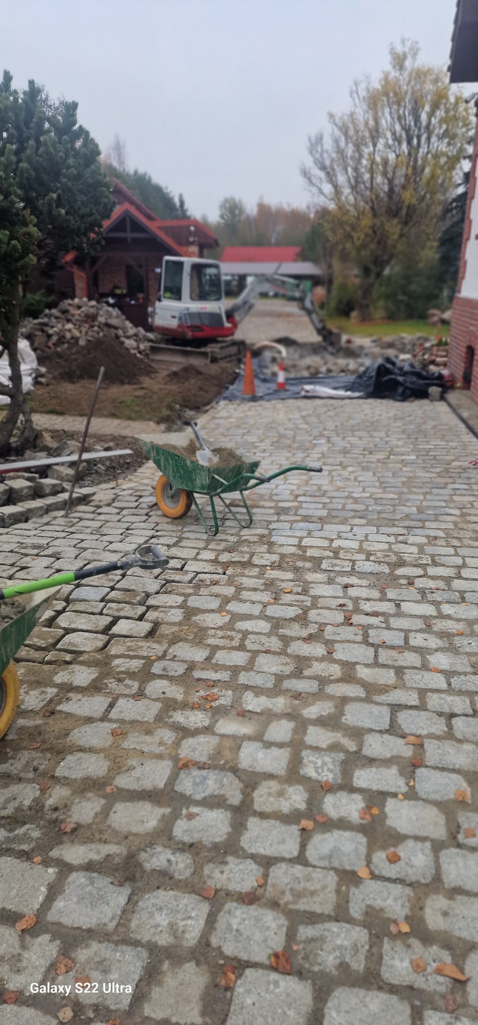 Układanie kostki brukowej z granitu na posesji, widoczna koparka, taczki i narzędzia, w tle budynki.