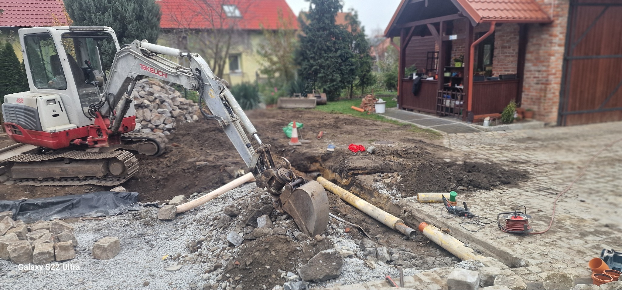 Mini koparka Takeuchi B235 przy wykopie z ułożonymi rurami kanalizacyjnymi, widoczny kabel zasilający, kostka brukowa i budynek gospodarczy w tle.