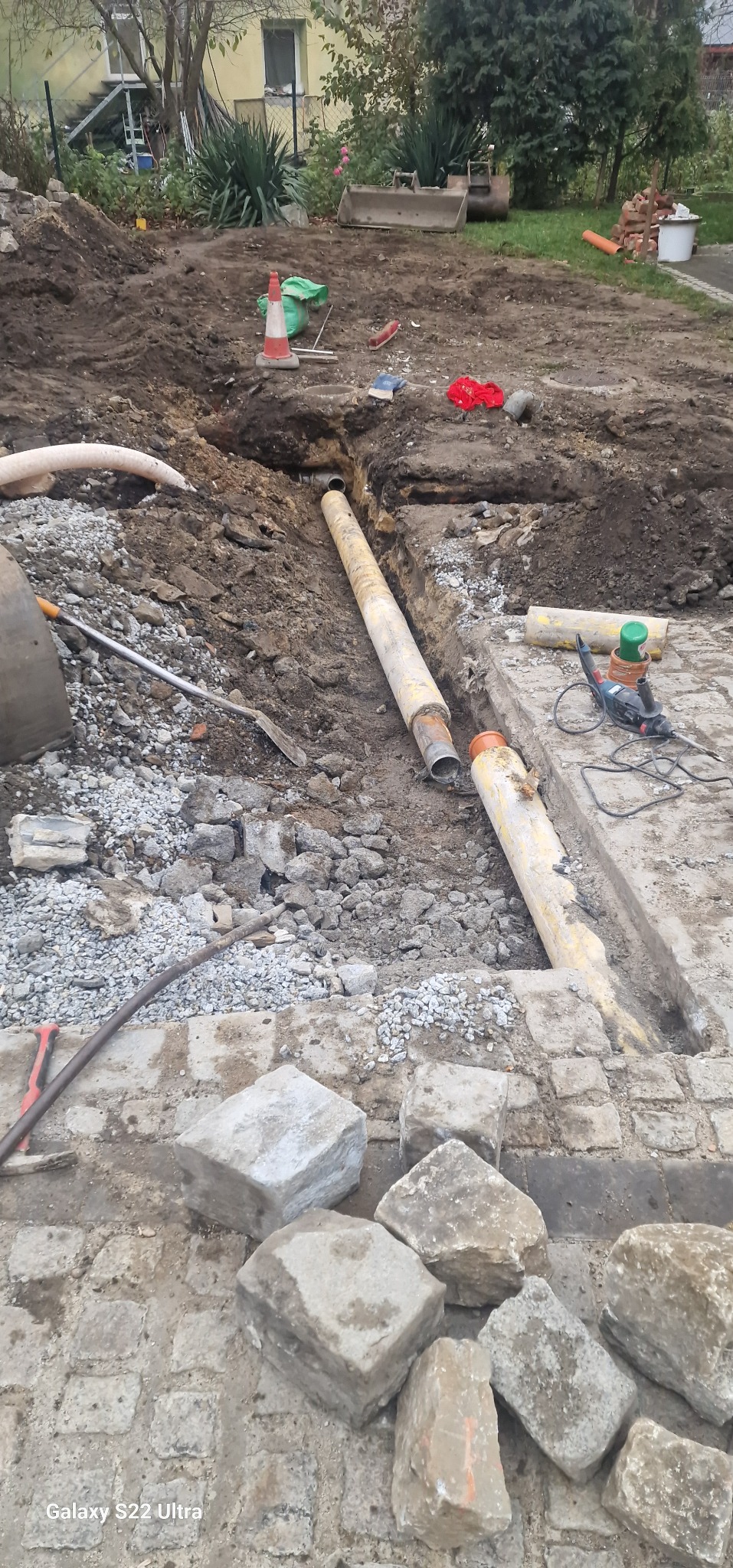 Prace ziemne związane z układaniem rur kanalizacyjnych w wykopie, widoczne narzędzia, fragment domu w tle.