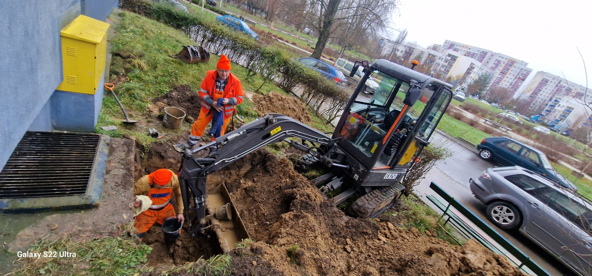 Dwaj pracownicy w pomarańczowych kombinezonach pracują w wykopie obok budynku, używając małej koparki do prac ziemnych, z widoczną żółtą skrzynką gazową i budynkami mieszkalnymi w tle.