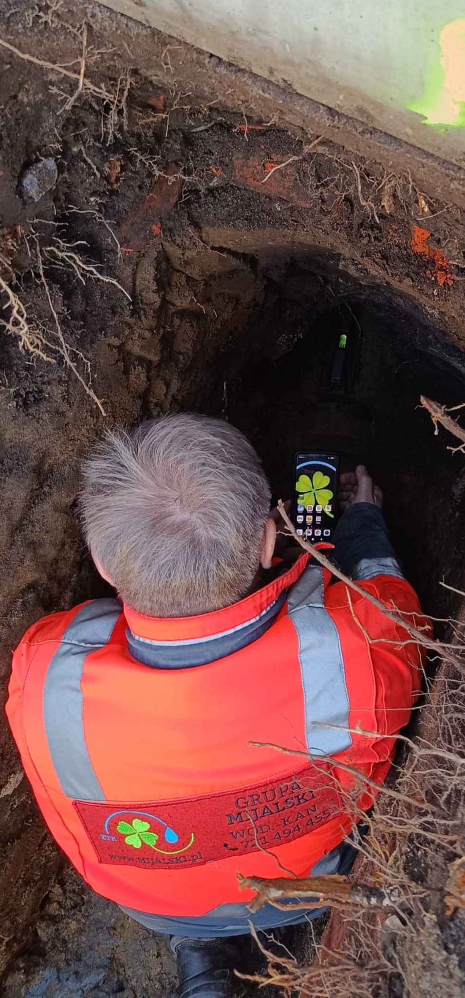 Pracownik w pomarańczowej kamizelce z logo firmy robi zdjęcie telefonem w wykopie z widoczną rurą i korzeniami drzew
