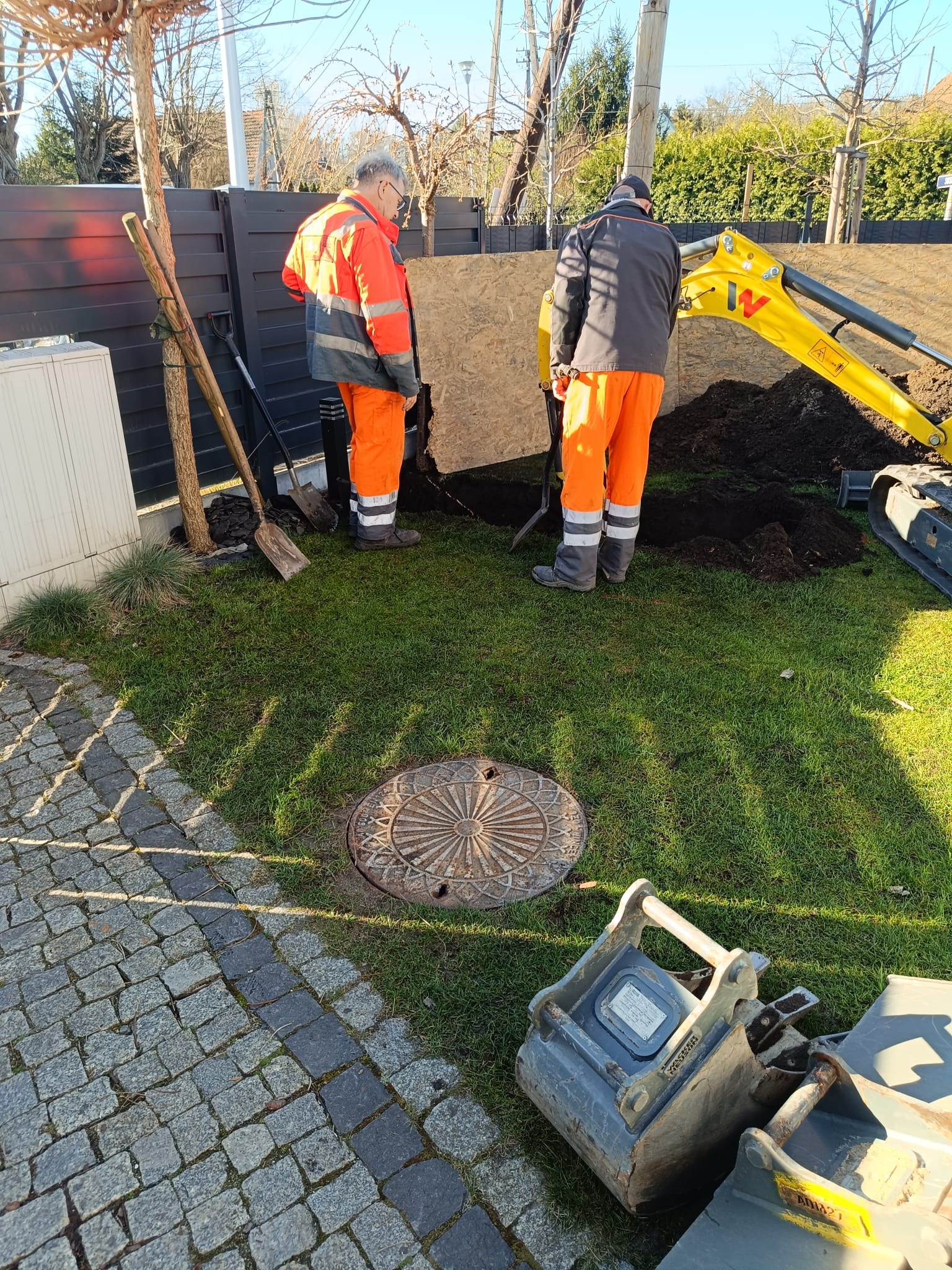 Dwaj pracownicy w odblaskowych ubraniach, koparka i wykopana ziemia na trawniku, widoczna studzienka kanalizacyjna, prawdopodobnie przygotowanie do podłączenia kanalizacji lub budowy szamba.