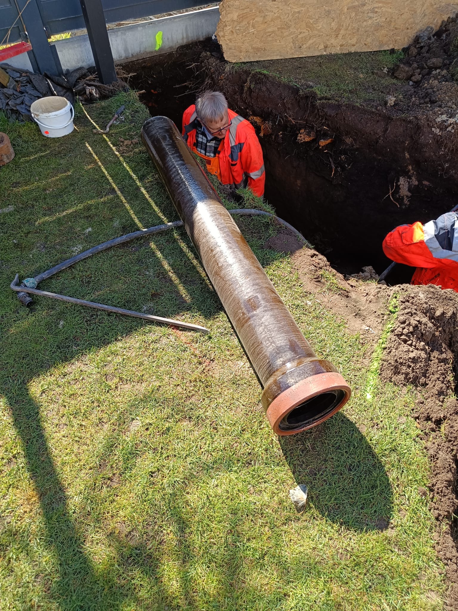 Przygotowanie do ułożenia rury kanalizacyjnej z uszczelką na trawie obok wykopu, pracownik w pomarańczowym kombinezonie w dole wykopu.