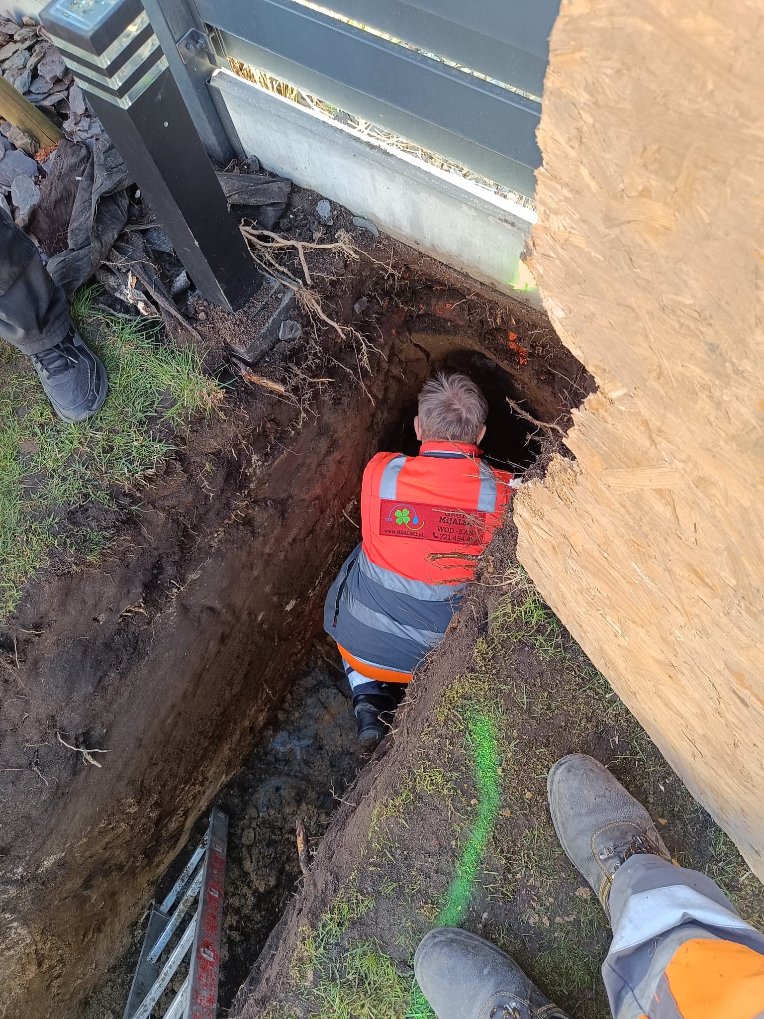 Pracownik w pomarańczowej kamizelce z logo firmy wod-kan w głębokim wykopie ziemnym, widoczna drabina, fragment ogrodzenia i zabezpieczenie wykopu płytą OSB. Zaznaczona linia wykopu zielonym laserem.