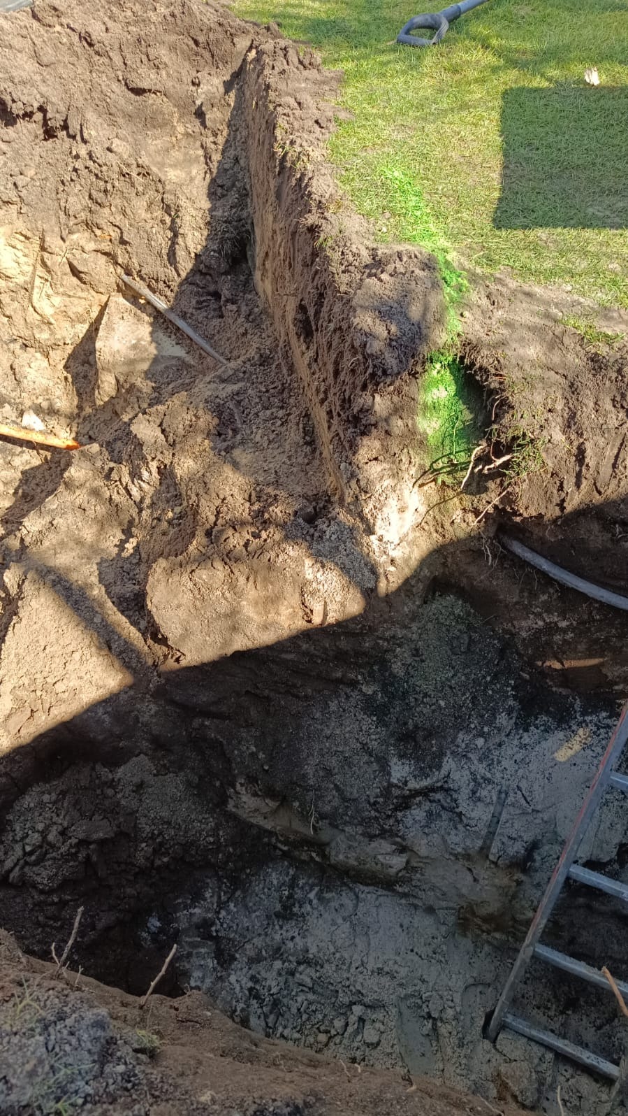Wykop w ziemi z widocznymi rurami, drabina oparta o ścianę wykopu, trawa w tle, widoczne ślady zielonej farby.