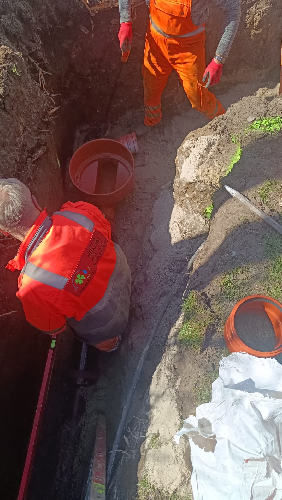 Pracownicy w pomarańczowych kombinezonach w wykopie podczas montażu rur kanalizacyjnych; widoczny fragment rury, poziomica i elementy szalunku