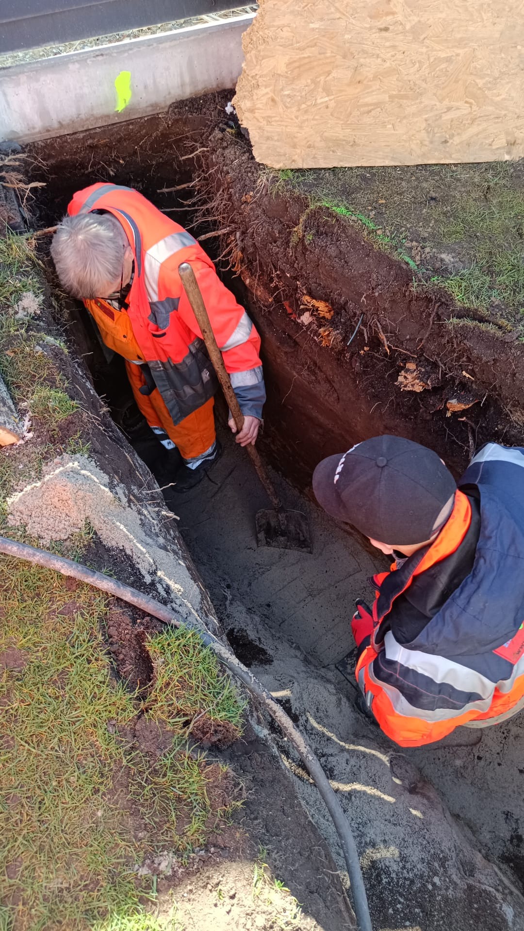 Dwóch pracowników w odblaskowych ubraniach kopie dół w ziemi, widoczne korzenie i rury, jeden z nich używa łopaty.