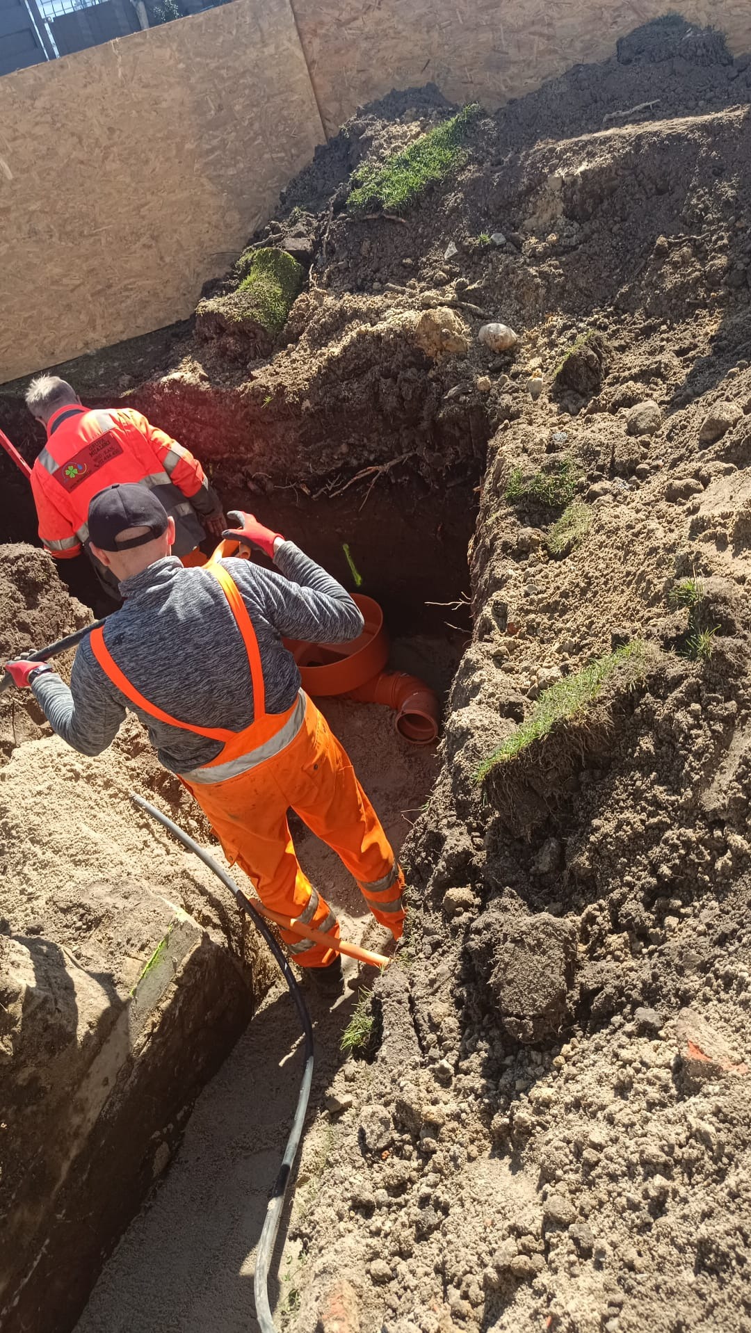 Dwóch pracowników w pomarańczowych kombinezonach układa pomarańczowe rury kanalizacyjne w wykopie ziemnym, widok z góry na plac budowy.