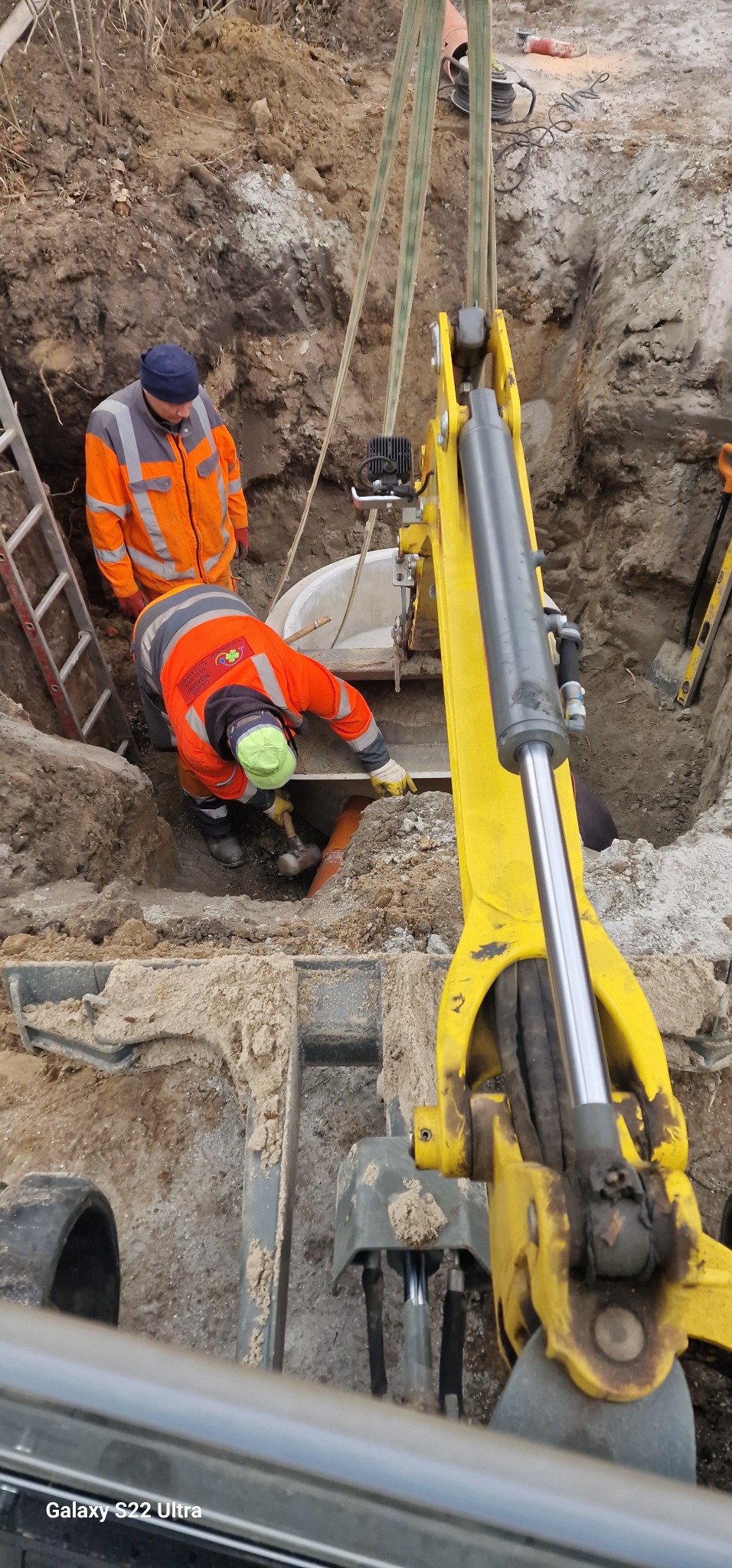 Montaż betonowego kręgu studni przez dwóch pracowników w pomarańczowych uniformach, z użyciem małej koparki w wykopie ziemnym, widok z góry.