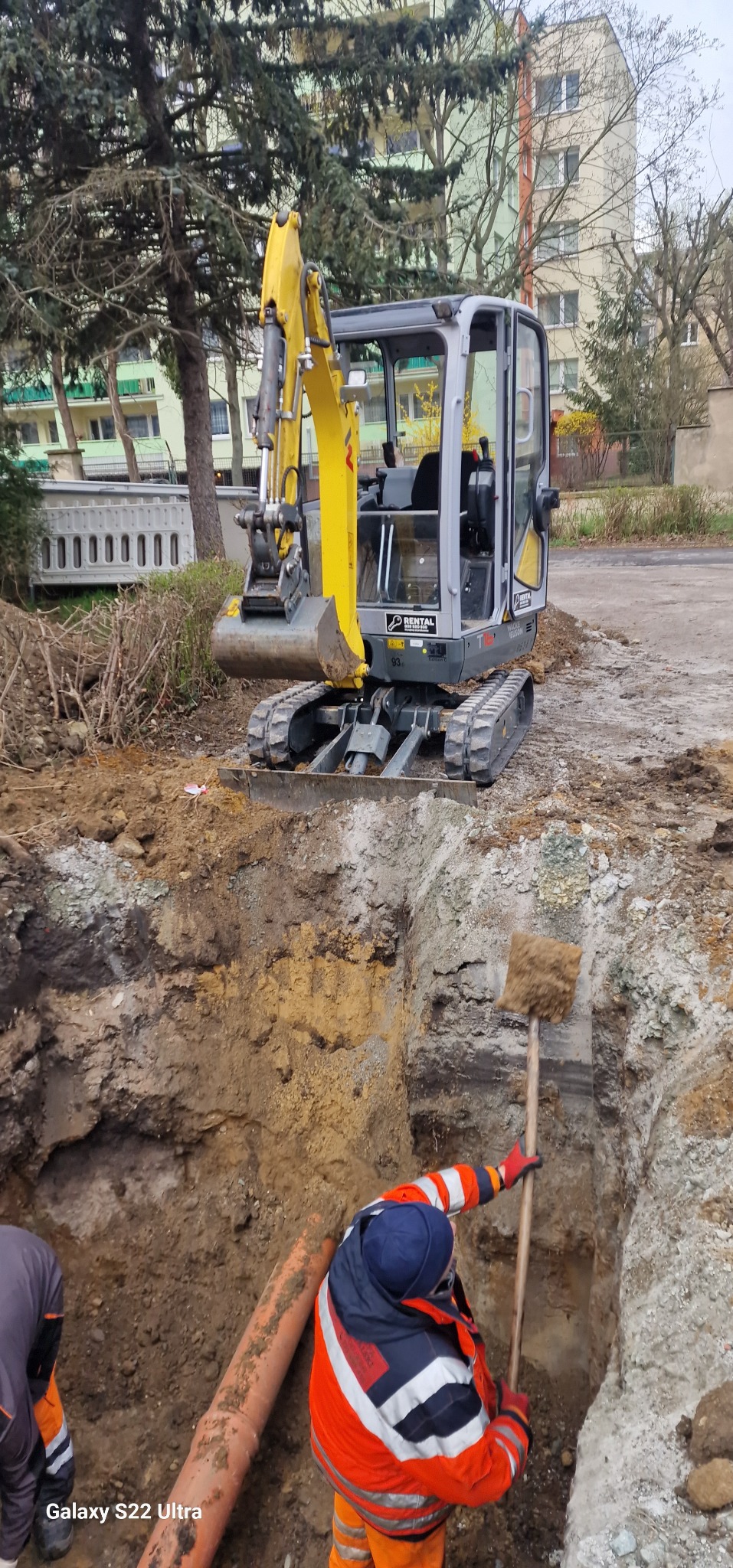 Prace ziemne przy wykopie pod rurę kanalizacyjną, widoczny pracownik z łopatą w pomarańczowej kurtce oraz minikoparka na powierzchni, w tle budynki mieszkalne.