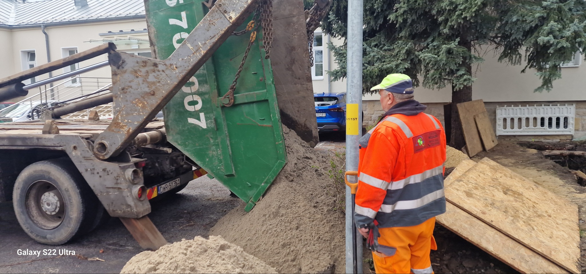 Wyładunek piasku z kontenera hakowego na placu budowy przyłącza wodno-kanalizacyjnego, pracownik w pomarańczowym ubraniu roboczym nadzoruje proces, widoczny wykop zabezpieczony płytami OSB, w tle...