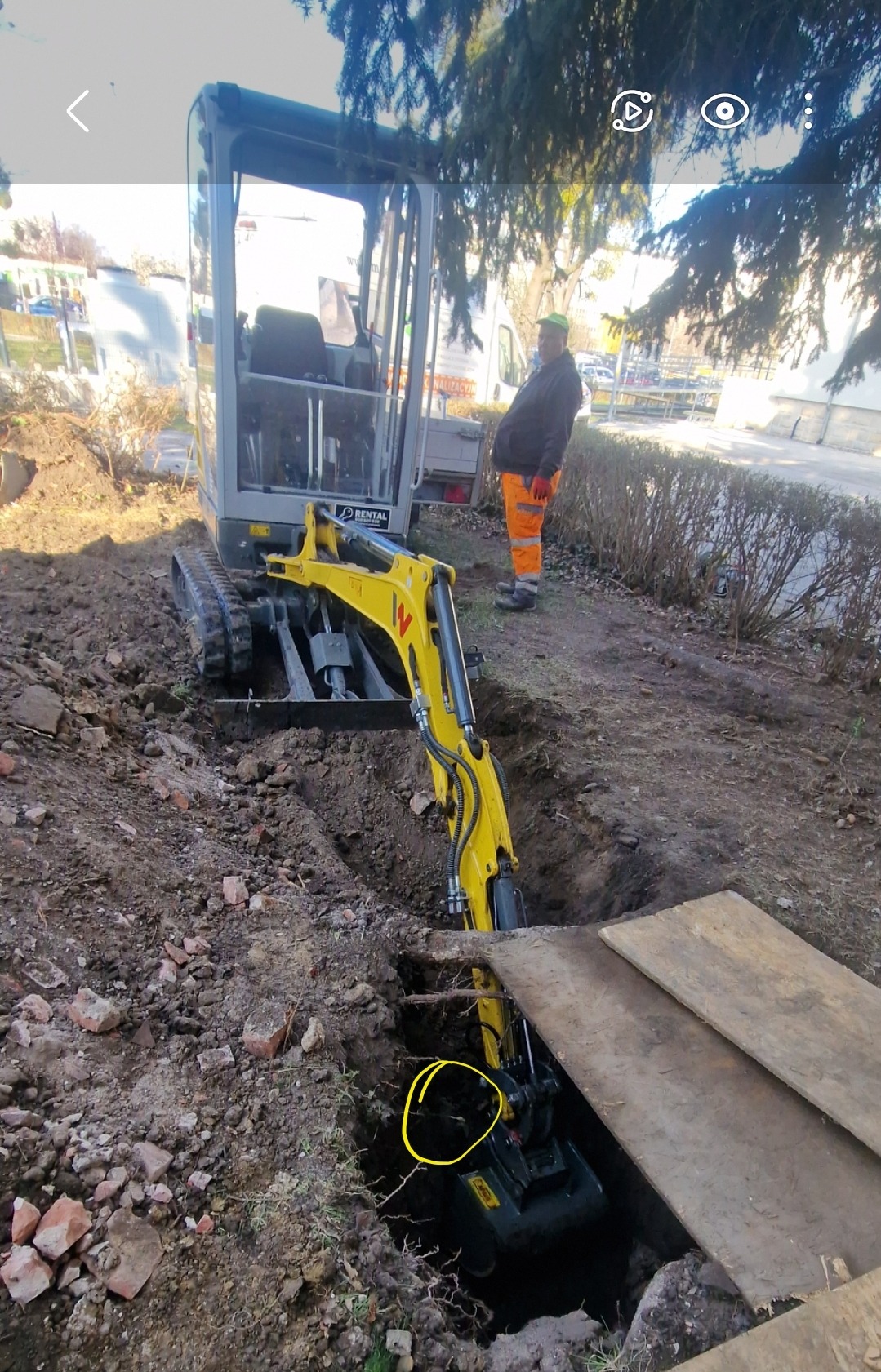 Żółta mini koparka zagłębia się w wykop w ziemi, obok stoi pracownik w pomarańczowych spodniach i zielonej czapce, widoczna tablica RENTAL na koparce.