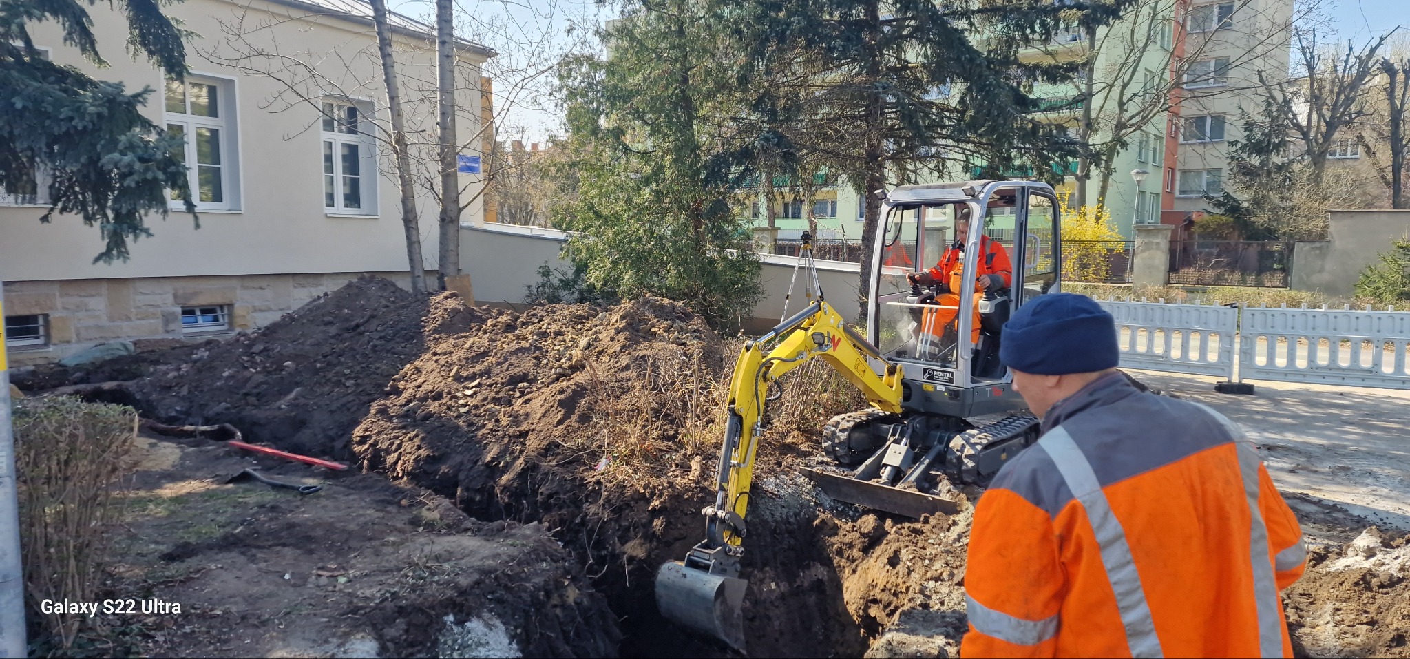 Żółta mini koparka podczas wykopu ziemi pod instalację, widoczny pracownik w kabinie oraz drugi pracownik w pomarańczowej kurtce z tyłu, w tle budynek i drzewa.