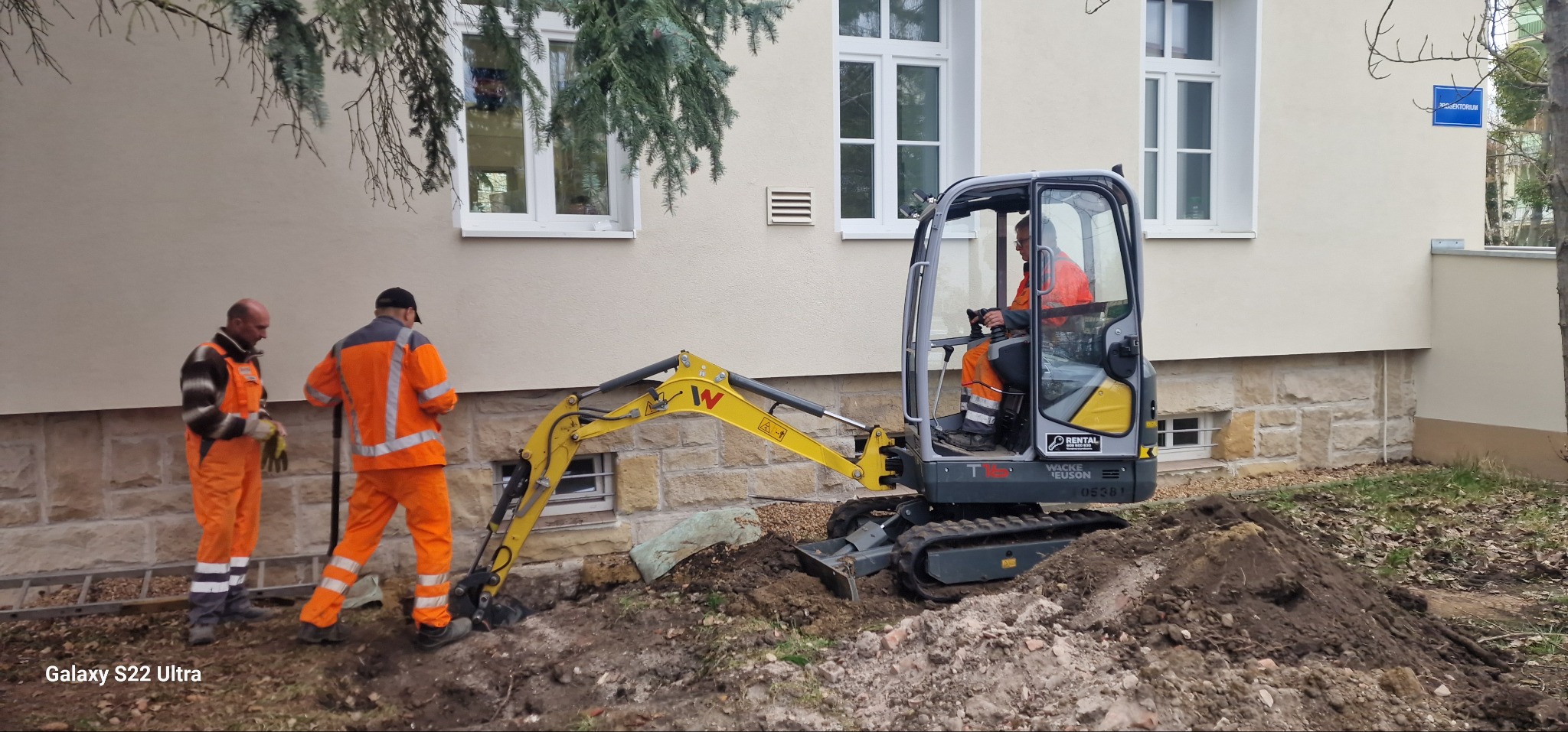 Prace ziemne przy budynku: operator w kabinie żółtej mini-koparki Wacker Neuson, dwóch robotników w pomarańczowych kombinezonach obok wykopu, budynek z oknami w tle.