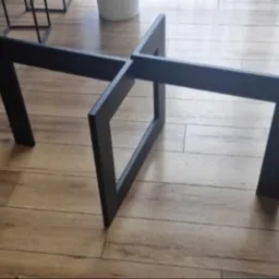 Czarna, geometryczna podstawa stolika kawowego z metalowych profili na drewnianej podłodze. Minimalistyczny design.