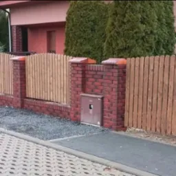 Nowoczesne ogrodzenie drewniane z ceglanymi słupkami i podmurówką, oddzielające posesję od chodnika z kostki. Widoczne elementy architektury domu w tle.