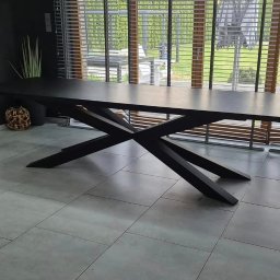 cayetann welding - Stół z czarnym blatem i geometryczną podstawą w kształcie litery X, na tle okna z żaluzjami. Minimalistyczny design, szare płytki podłogowe.