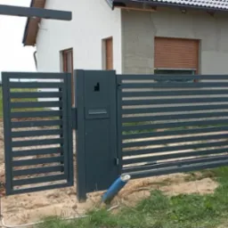 Nowoczesna, pozioma brama wjazdowa i ogrodzenie panelowe w kolorze antracytowym. Brama uchylona, widoczny fragment domu w tle. Słupek oświetleniowy.