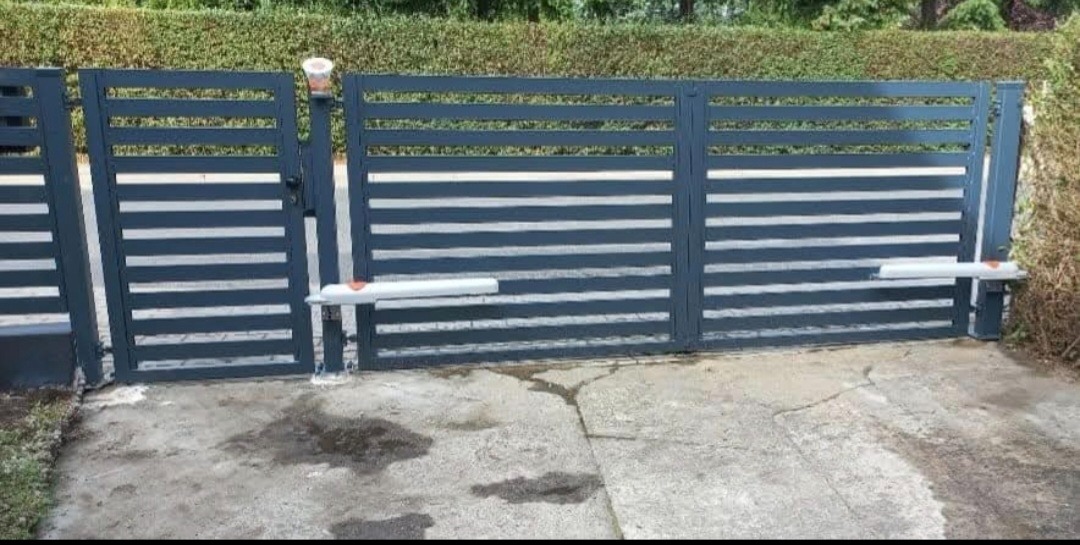 Nowoczesna, pozioma brama wjazdowa z automatyką, pomalowana na grafitowo, na tle zielonego żywopłotu. Widoczne siłowniki bramy.