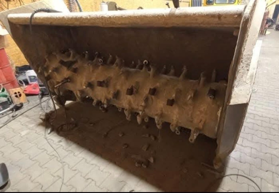 Metalowy, brudny frez do gleby z licznymi zębami, wewnątrz metalowej obudowy. Urządzenie stoi na posadzce z kablami w tle. Widoczne ślady użytkowania.