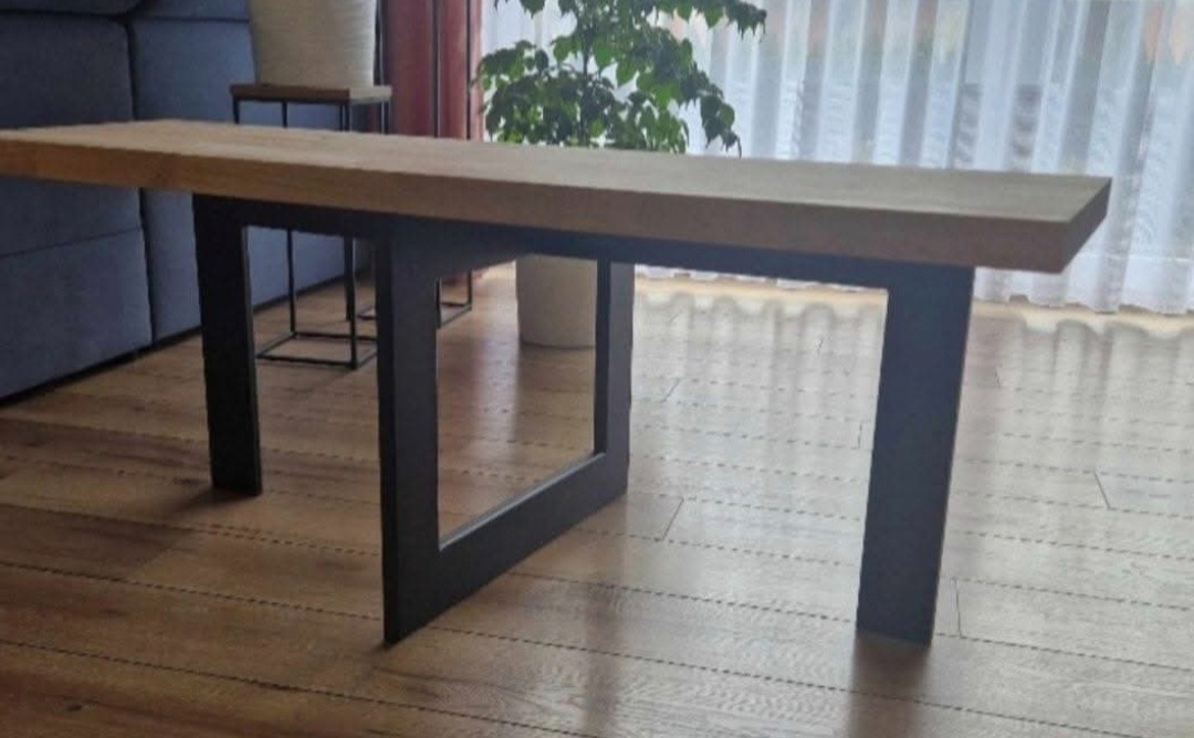 Stół z blatem z jasnego drewna i czarnymi, metalowymi nogami o nowoczesnym designie. W tle widoczny fragment sofy i okno z firanką.